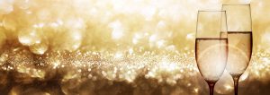 Two champagne glasses filled with bubbling champagne stand side by side against a sparkling gold, festive background with bokeh lights.