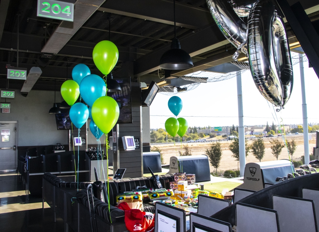 A decorated golf bay features blue and green balloons, large silver 30 balloons, and a table with snacks and gifts, suggesting a 30th birthday celebration at an indoor golf venue, with numbered bays visible.