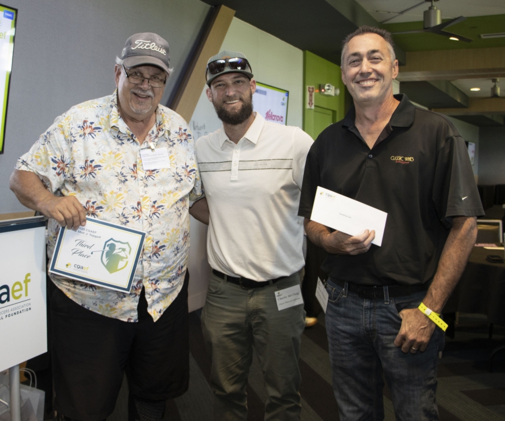 Three men pose and smile at an indoor event; one holds a certificate, another holds an envelope. A sign reads cgaef. Sponsor logos are visible on the wall behind them. Other attendees sit at tables in the background.