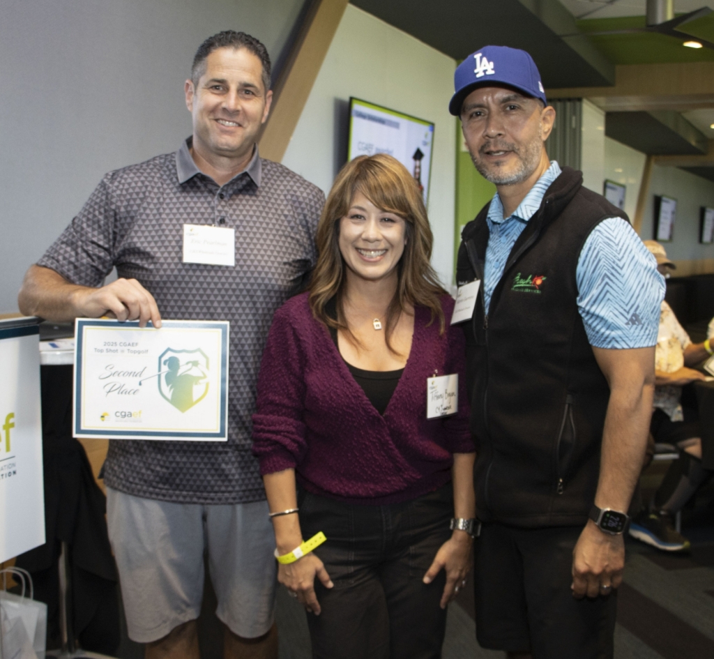 Three people smiling at the camera; one holds a certificate reading Second Place. They stand in a conference room with attendees seated at tables. A CGAEF banner and a screen displaying a woman are visible in the background.