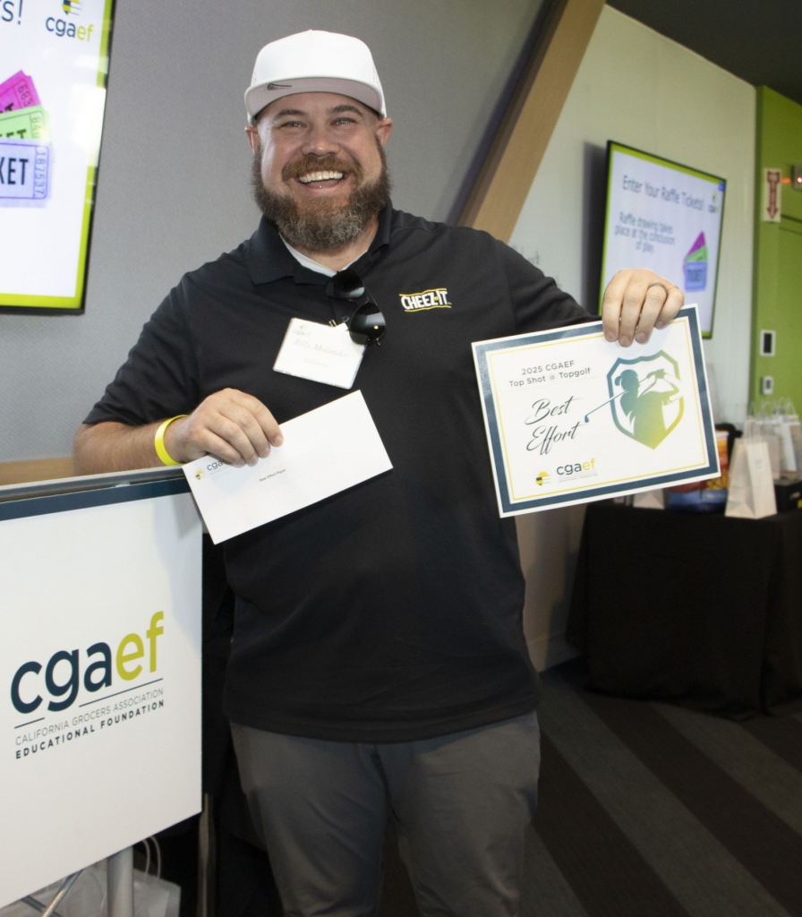 A smiling man in a black shirt and cap holds an envelope and a Best Effort certificate. Behind him is a raffle sign and a screen with ticket information at a CGAEF event.
