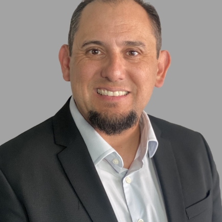 A man with short dark hair and a goatee, wearing a black suit jacket and white shirt, smiles at the camera against a plain gray background.