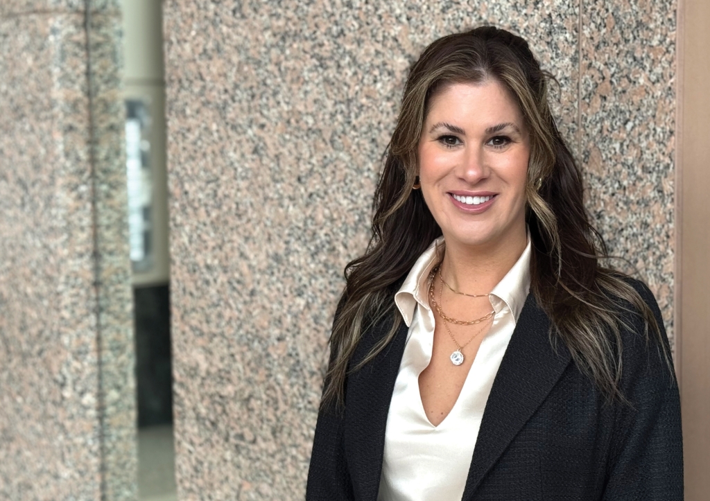 A woman with long, wavy brown hair wearing a black blazer and cream blouse stands smiling in front of a granite wall.