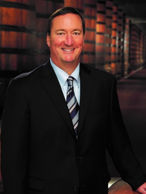 Smiling man in a suit standing by barrels in a dimly lit cellar or warehouse.