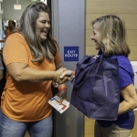 Two women smile as they shake hands; one holds tickets, the other carries a large blue bag. They stand near a door with an Exit Route sign, suggesting a friendly interaction in a public setting.