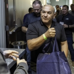 A man in a black shirt smiles while holding a blue tote bag, standing in a hallway among a group of people. One person in the foreground gestures toward him.