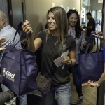 A smiling woman hands out blue tote bags with a CGAEF logo to people in a doorway. Another woman behind her also holds a bag, and several people are in the background. The setting appears to be indoors at an event.