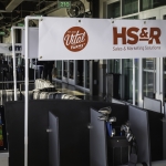 A display banner with the logos of Vital Farms and HS&R Sales & Marketing Solutions hangs above golf club bags at an indoor golf facility. The setting includes golf bays, striped chairs, and glass windows.
