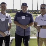 Three men in golf attire stand smiling at a driving range, each holding a golf club. Two wear name badges, and the background shows a sunny day with green golf bays and distant trees.