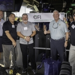 Five smiling men holding golf clubs stand together indoors at a golf venue, posing in front of an OFI Organic Foods International sign. Golf bays and screens are visible in the background.