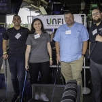 Five people stand indoors holding golf clubs, smiling at the camera. They wear name tags and casual clothes. A UNFI sign is visible behind them, and the setting suggests an indoor golf or event venue.