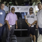 Four men stand smiling in front of a Treasury Americas sign at an indoor golf venue. They are casually dressed and holding golf clubs, with equipment and other people visible in the background.