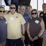 Six adults stand together and smile for a group photo at an outdoor event. Most are wearing sunglasses and name badges. They are casually dressed, and the background shows a covered, open-air venue with sunlight streaming in.