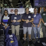 Six people stand side by side indoors at a golf venue, posing with golf clubs and wearing nametags. They are smiling, and behind them is a banner and golf equipment.