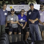 Five men stand indoors holding golf clubs, smiling at the camera. They wear name tags and casual golf attire. A Molson Coors Beverage Company sign is visible behind them.