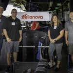 Four people stand smiling with golf clubs at an indoor driving range in front of a sign that reads Kellanova. Golf bags and driving range stalls are visible in the background.