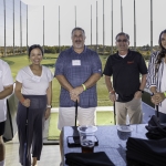 Five people stand indoors at a golf driving range, holding golf clubs and smiling at the camera. The background shows golf bays, netting, and an outdoor field under clear daylight.