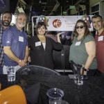 Five people stand and smile in front of a “Wholesale Curers” banner at an indoor event. Halloween-themed decorations and empty cups are visible on the table in front of them.