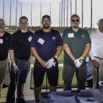 Five men stand side by side at an indoor golf driving range, all smiling at the camera; three are holding golf clubs, and all are wearing name tags and casual attire. Golf nets and a clear sky are visible in the background.