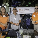 Three people stand and smile near a “TruGrocer Federal Credit Union” sign at an indoor event. Two wear orange shirts and name tags; one is in a gray polo. Drinks and bags are on the table in front of them.