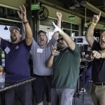 Four men enthusiastically cheer with raised arms inside a sports bar or entertainment venue, drinks on the table, and sports screens in the background.