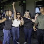 Four people stand together indoors, smiling and posing with their arms outstretched in excitement. They wear casual clothes and name tags, suggesting a fun group or corporate event. Other people and tables are visible in the background.