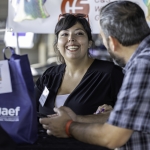 A smiling woman sits at a table speaking with a man in a plaid shirt. There are bags and a name badge on the table, and a banner with partially visible text is in the background.
