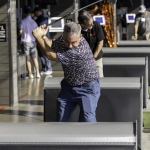 A man in a patterned shirt and blue pants prepares to swing a golf club at an indoor driving range, with other people and golf bays visible in the background.