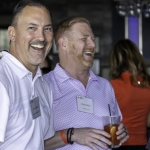Two men wearing name tags laugh together at an indoor event. One man holds a drink with a straw and an orange slice. Other people are visible in the background, but out of focus.