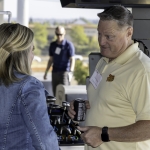 A man in a pale yellow polo shirt holds a can and talks to a woman in a denim jacket at an outdoor event, with golf clubs and another person blurred in the background.