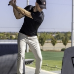 A man in a black cap and shirt swings a golf club at a driving range, preparing to hit a golf ball on a tee, with trees and a clear blue sky in the background.