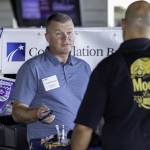 A man in a blue shirt with a name tag speaks to another person at an indoor event booth. The booth has a banner in the background and various items on the table, including beads and small objects.