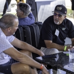 Two men sit on a black couch having a conversation at an indoor golf venue; one wears a cap and sunglasses, and the other has a wristband. Drinks and phones are on the table in front of them. Two more people sit in the background.