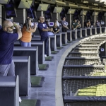 People stand in individual bays at a multi-level driving range, some watching and others swinging golf clubs. Monitors are mounted above each bay, and a safety net runs along the open edge. The atmosphere appears lively.