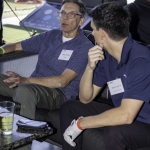 Two men sit on a black couch, engaged in conversation at an indoor golf venue. Both wear name tags, and one man has a drink on the table beside him. Golf equipment and a Honda sign are visible in the background.