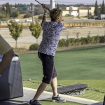A person wearing a patterned shirt, black shorts, and sneakers is swinging a golf club at a driving range, with trees, a road, and buildings visible in the background.