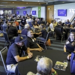 A large group of people sit at round tables in a modern, bright event room, engaging in conversation. Some look toward a speaker, while others talk in small groups. Event materials and tote bags are on tables and chairs.