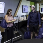 Five people stand and chat in a modern conference room, holding tote bags. Screens display event and scholarship information in the background. Some are casually dressed, and everyone is smiling or engaged in conversation.