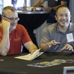 Two men sit at a round table during a meeting. One wears a red polo and glasses, looking thoughtful, while the other in a gray polo smiles and gestures. Papers and a glass of water are on the table.