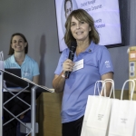 A woman in a blue shirt speaks into a microphone at a podium, smiling, while another woman stands nearby. White gift bags and a cardboard box are visible in the foreground. A screen displays a photo and text in the background.