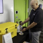A man smiles while arranging a figurine on a table next to a large yellow cooler and a white gift bag. There are chairs and another person in the background of the room.