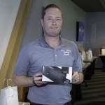 A man in a light blue polo shirt holds a boxed Bose speaker and stands indoors. Gift bags and a table are visible in the background.