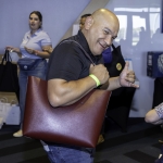 A smiling man poses with a maroon shoulder bag, making a shaka hand gesture. Other people and tables with bags are visible in the background at an indoor event.