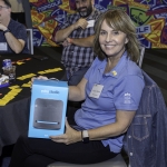 A smiling woman in a blue shirt sits at a table holding an Echo Studio box. Other people are around the table with colorful Topgolf decor in the background.