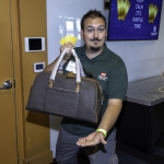 A man in a green shirt holds three yellow raffle tickets and a brown duffel bag, while gesturing with his other hand. A sign behind him reads, KEEP CALM ITS RAFFLE TIME.