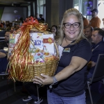 A smiling woman with long blonde hair and glasses holds a large gift basket with a red and gold ribbon. People are seated in the background at an indoor event.