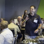 A man stands and smiles while others seated around him clap and smile in a conference room setting. The atmosphere is lively and positive, with food trays on a table in the foreground.