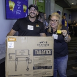 Two smiling people pose indoors, holding a large Traeger Tailgater grill box. The woman shows a yellow raffle ticket. In the background, a sign reads “Keep Calm It’s Raffle Time.” Other people are visible behind them.