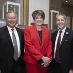 Three people in business attire smile and pose together at an indoor event; framed memorabilia and sports jerseys are displayed in the background.