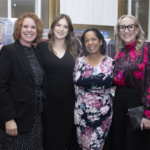 Four women pose together and smile at an indoor event, standing in front of tables displaying framed artwork and collectibles. They are dressed in business and floral attire.